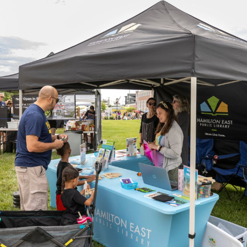 Outdoor library booth with a teal table labeled Hamilton East Public Library; staff assist visitors, including a man and children, under a black canopy.