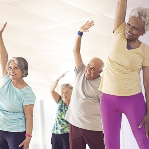 Group of seniors in a bright studio, standing with arms raised in a side stretch.