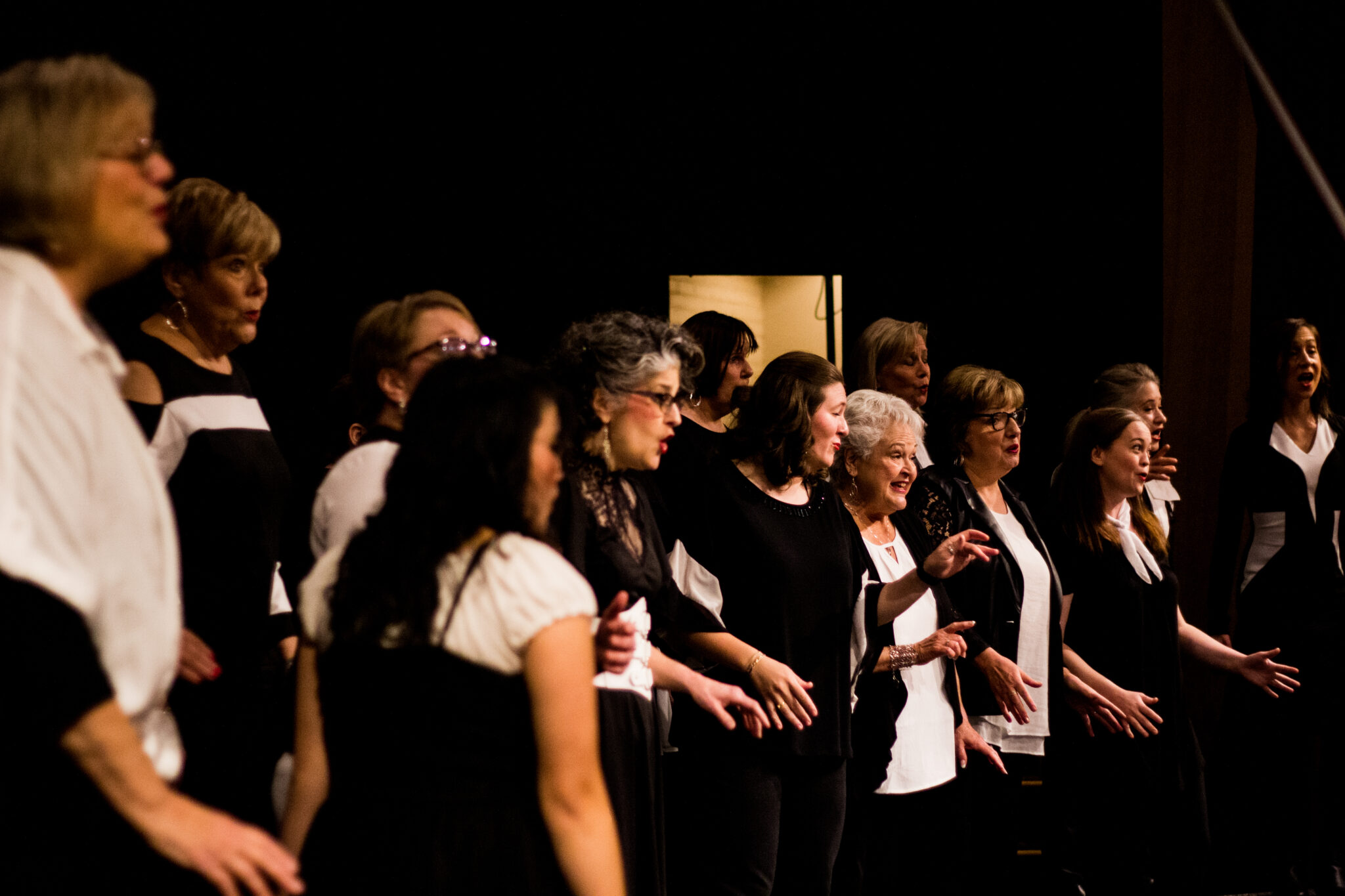 A choir of about 15, mostly women, singing on a dark stage in black and white outfits.