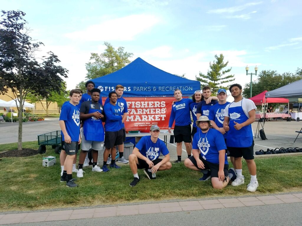 Group of youths in blue team shirts posing together in front of a Fisher's Farmers Market booth at an outdoor event.