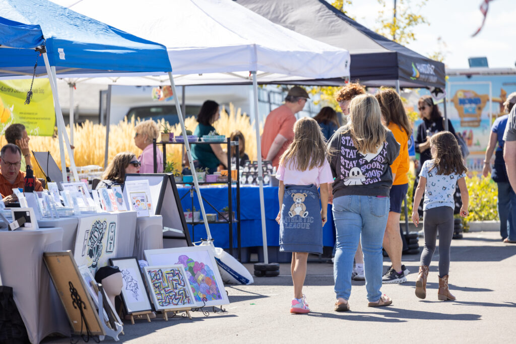 Outdoor market with blue and white tents; tables display colorful artwork as shoppers browse nearby.