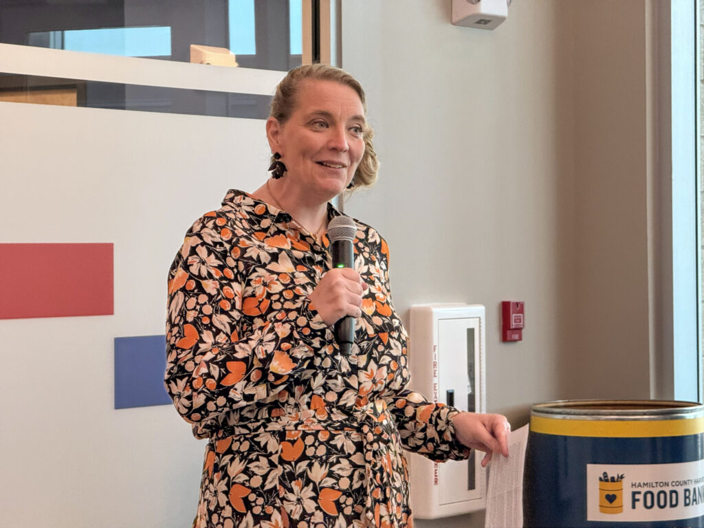 Woman speaking into a handheld microphone at a community event, wearing a orange–black floral dress, beside a food bank donation barrel.