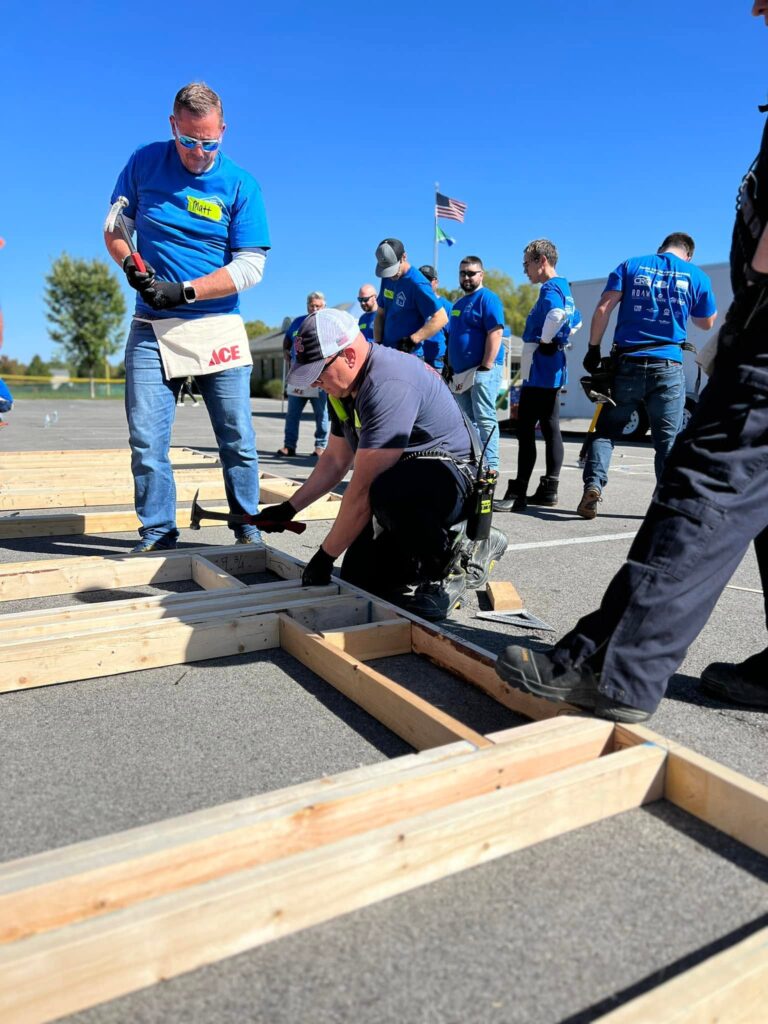 Group of men in blue shirts building wooden frames on a parking lot; one kneels with a hammer.