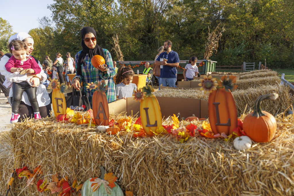 Group of people at a fall festival around hay-bale displays decorated with pumpkins, sunflowers, and autumn leaves; letters spell FALL in the foreground.