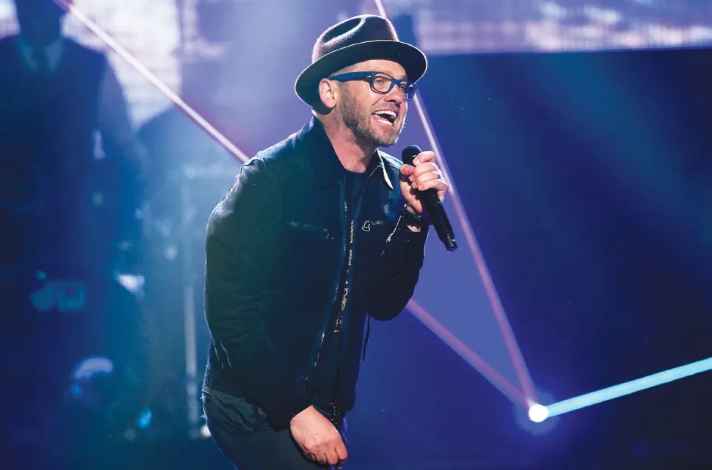 Male singer wearing a hat and glasses, performing on stage with a handheld microphone under blue lights.