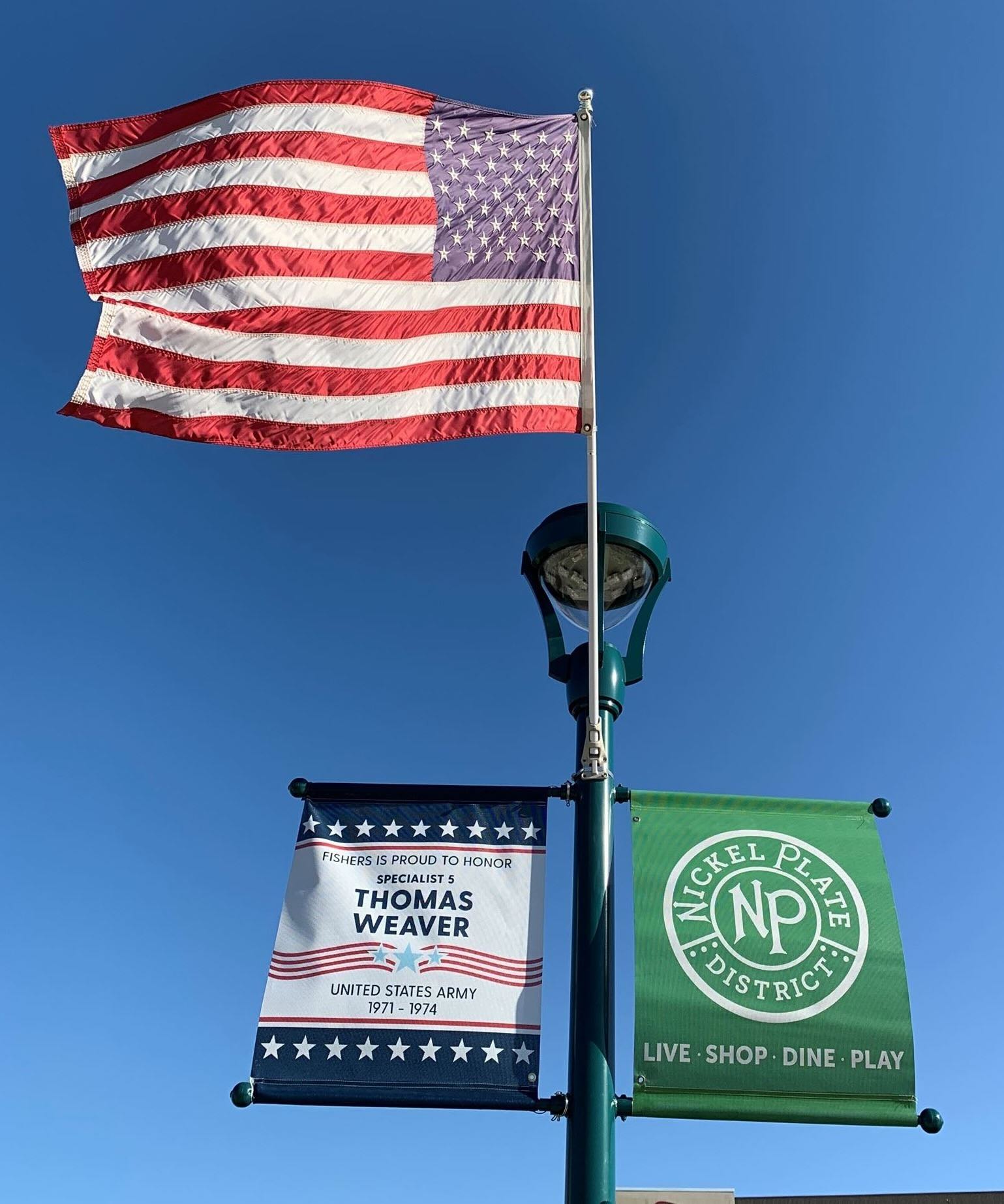 American flag on a flagpole with two banners reading Nickel Plate District and Thomas Weaver against a clear blue sky.