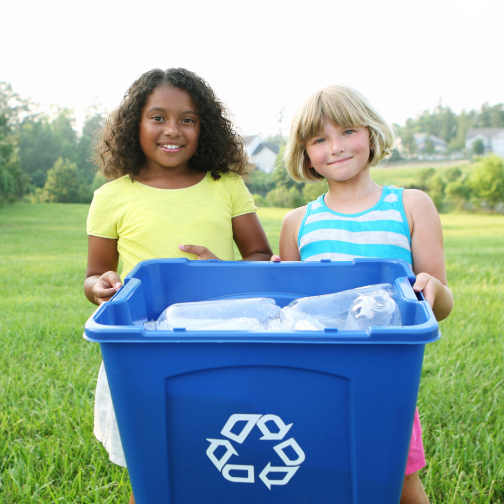 Two smiling girls stand in a grassy park behind a blue recycling bin filled with plastic bottles.